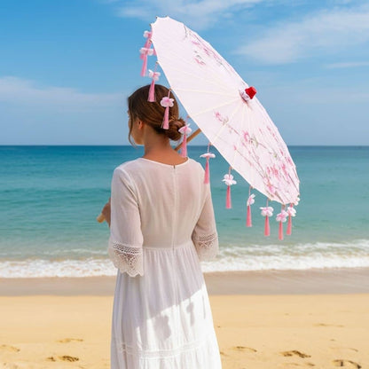 Femme en robe blanche tenant une ombrelle rose décorée de fleurs, face à la mer sous un ciel bleu