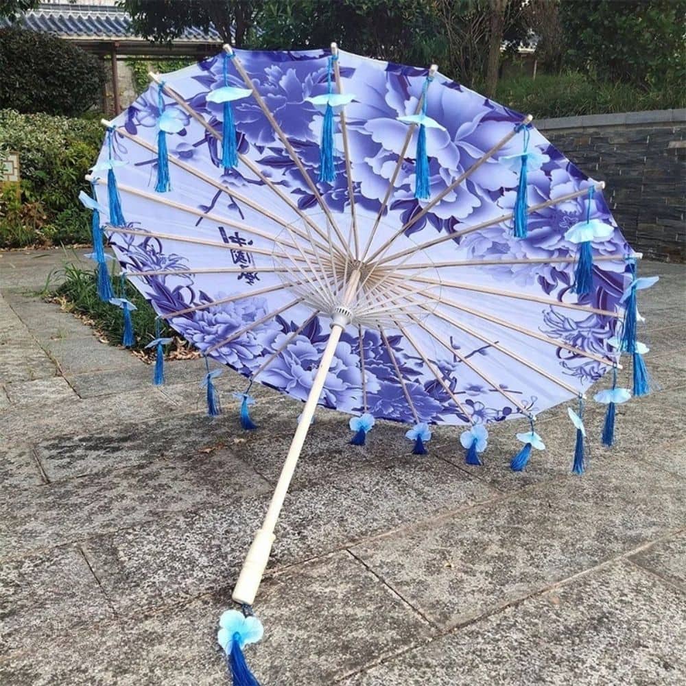 Ombrelle anti-UV violette ornée de motifs floraux, avec des franges bleues et un décor naturel en arrière-plan