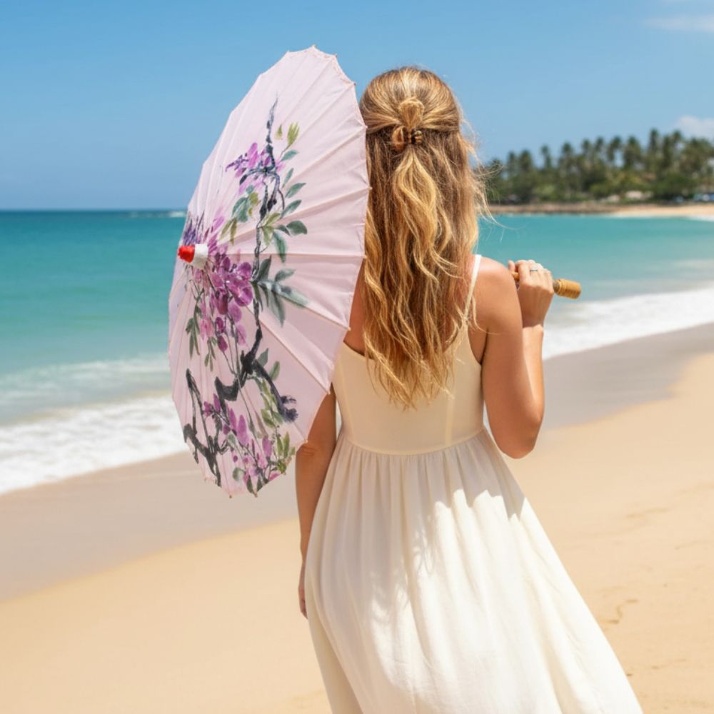 Femme en robe blanche tenant une ombrelle anti-UV violette ornée de fleurs, face à la mer turquoise