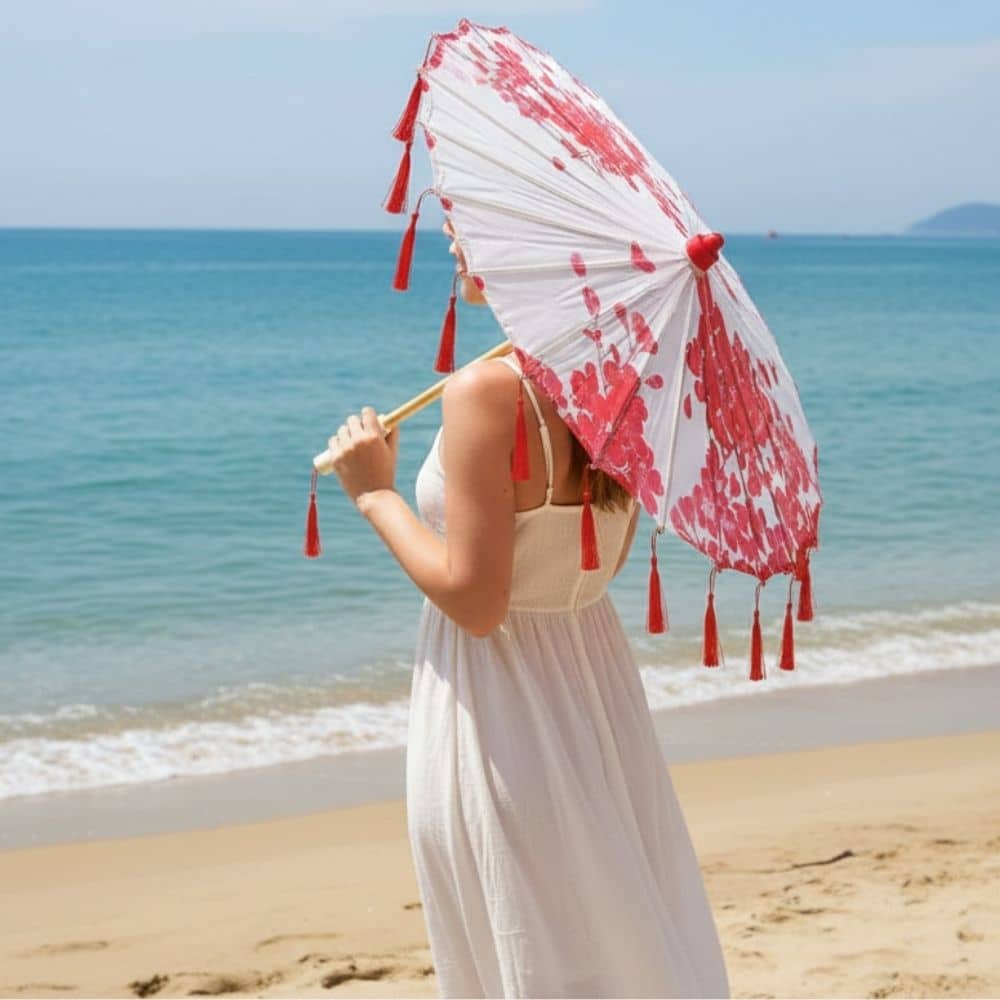 Femme en robe blanche tenant une ombrelle blanche ornée de motifs rouges sur une plage ensoleillée