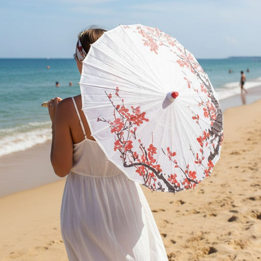 Femme en robe blanche tenant une ombrelle blanche ornée de fleurs rouges sur une plage ensoleillée