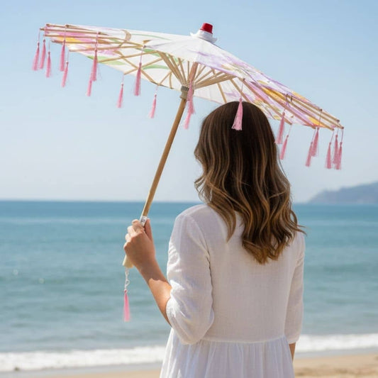 Une ombrelle blanche avec des franges roses, tenue par une femme en robe légère, sur une plage ensoleillée
