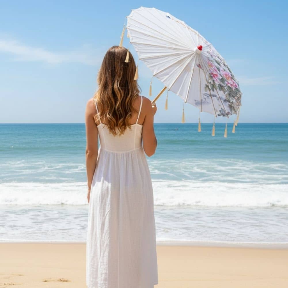 Femme en robe blanche tenant une ombrelle blanche décorée, face à l'océan sous un ciel bleu