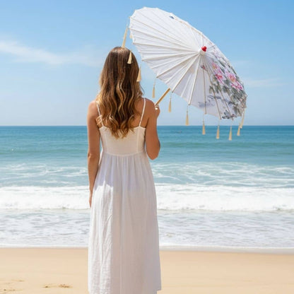 Femme en robe blanche tenant une ombrelle blanche décorée, face à l'océan sous un ciel bleu