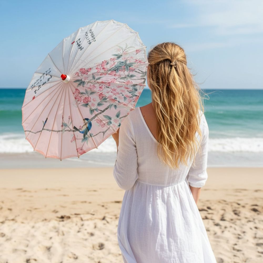 Femme portant une ombrelle rose décorée de fleurs, regardant la mer depuis une plage ensoleillée