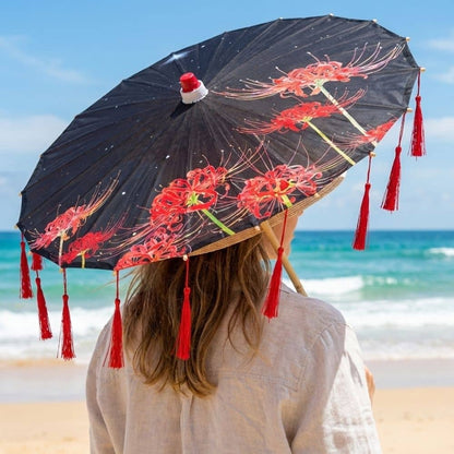 Femme vue de dos tenant une ombrelle noire ornée de fleurs rouges, avec des franges rouges, sur une plage ensoleillée