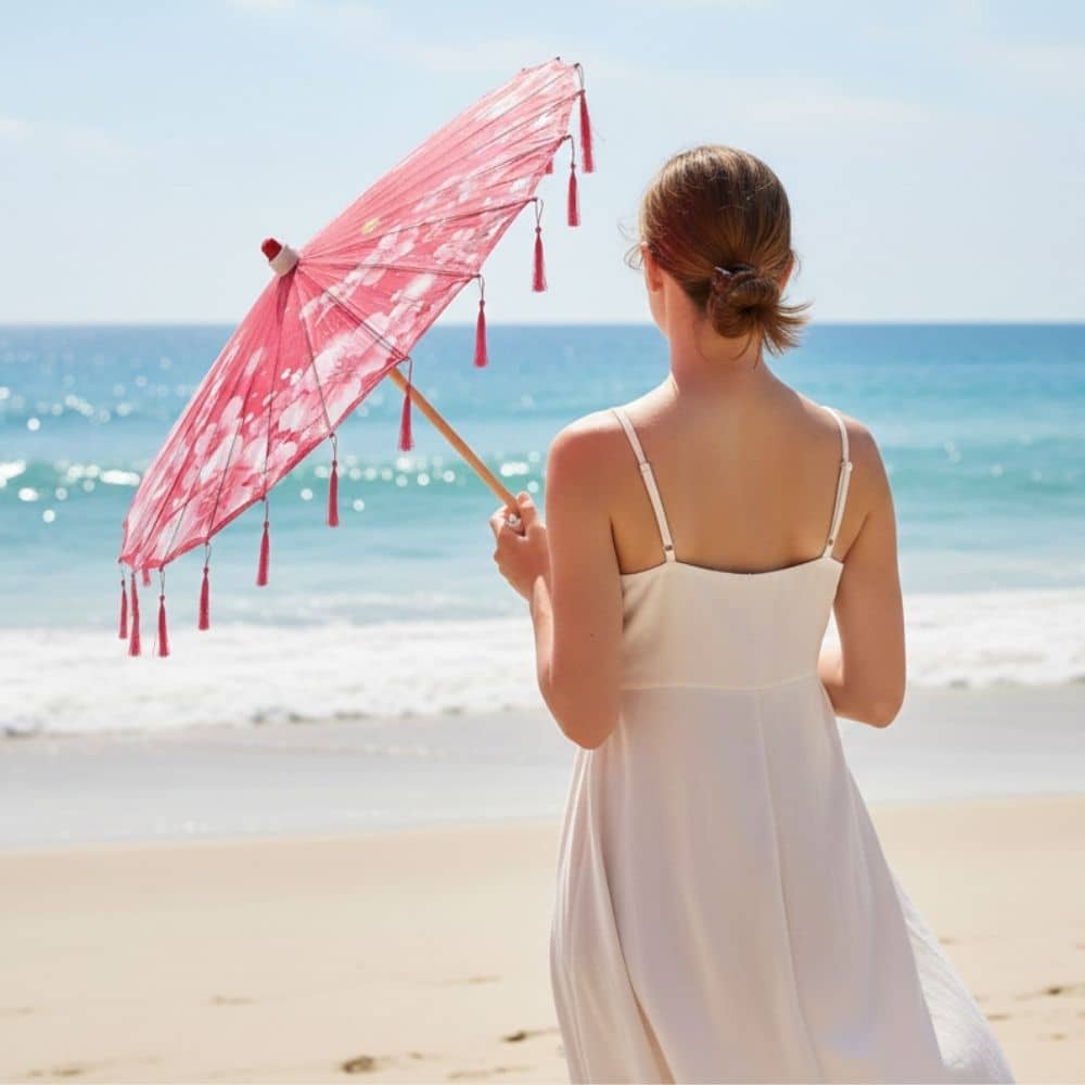 Femme de dos tenant une ombrelle fleurie rose, vêtue d'une robe blanche, face à la mer