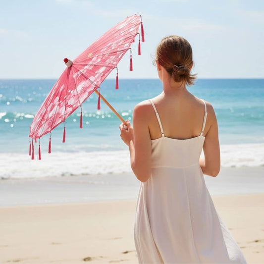 Femme de dos tenant une ombrelle fleurie rose, vêtue d'une robe blanche, face à la mer