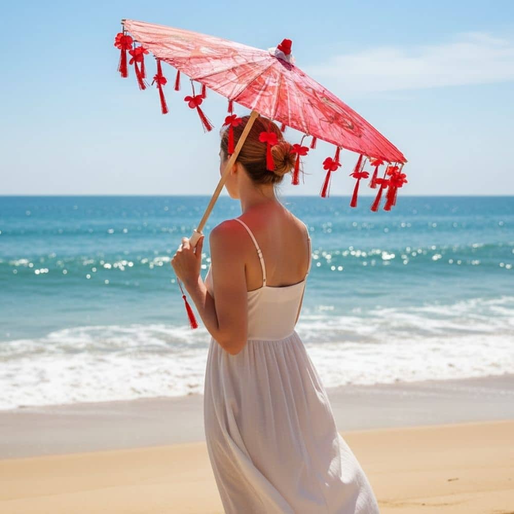 Femme en robe blanche tenant une ombrelle japonaise rouge ornée de franges, face à la mer ensoleillée