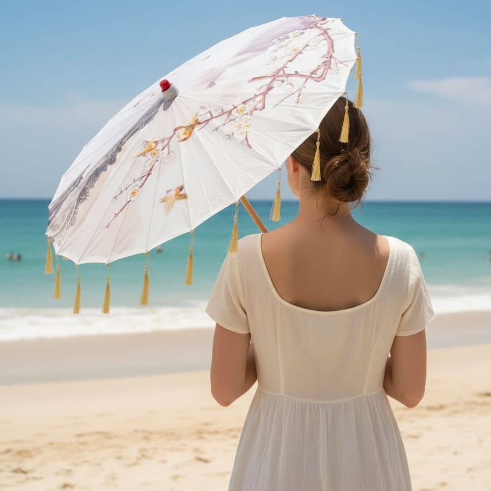 Femme de dos portant une robe blanche, tenant une ombrelle japonaise ornée de motifs floraux sur une plage ensoleillée