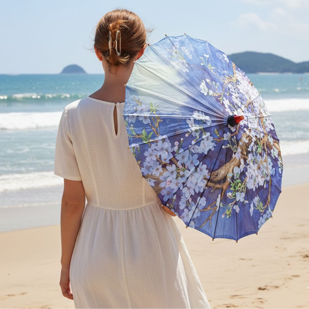 Femme de dos portant une ombrelle japonaise bleue ornée de fleurs blanches, sur une plage ensoleillée