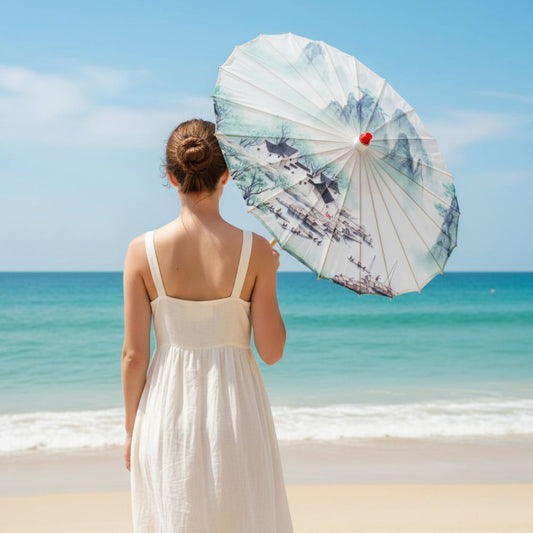 Femme en robe blanche tenant une ombrelle japonaise décorée, face à une mer turquoise sous un ciel bleu