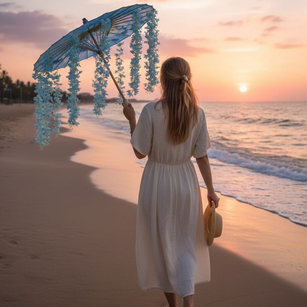 Femme en robe blanche tenant une ombrelle bleue décorée, marchant sur la plage au coucher du soleil
