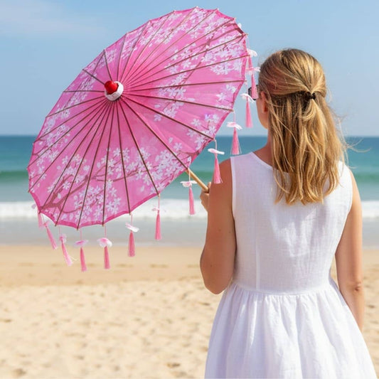 Une ombrelle pliante rose avec des motifs floraux et des pompons, tenue par une femme sur la plage