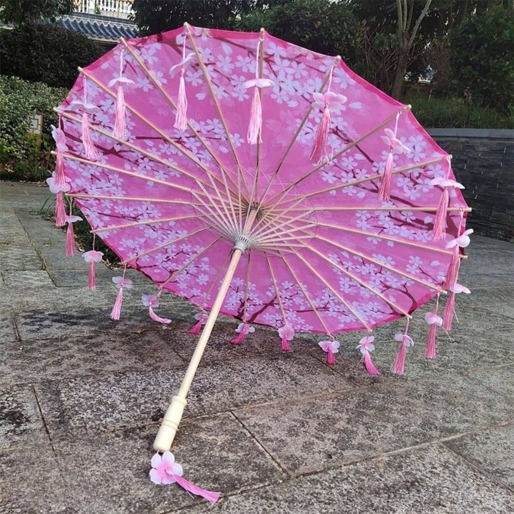 Ombrelle pliante rose avec motifs floraux et franges, posée sur un sol en pierre, entourée de verdure