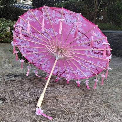 Ombrelle pliante rose avec motifs floraux et franges, posée sur un sol en pierre, entourée de verdure