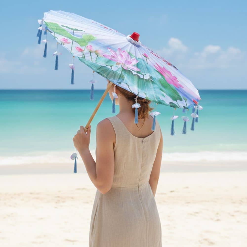 Femme debout sur la plage tenant une ombrelle pliante aux motifs floraux, sous un ciel bleu clair