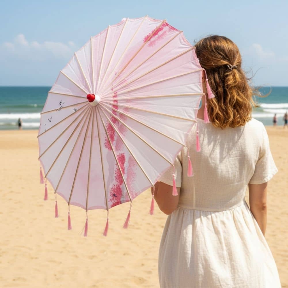 Femme portant une ombrelle rose à motifs floraux, sur une plage ensoleillée avec des vagues en arrière-plan