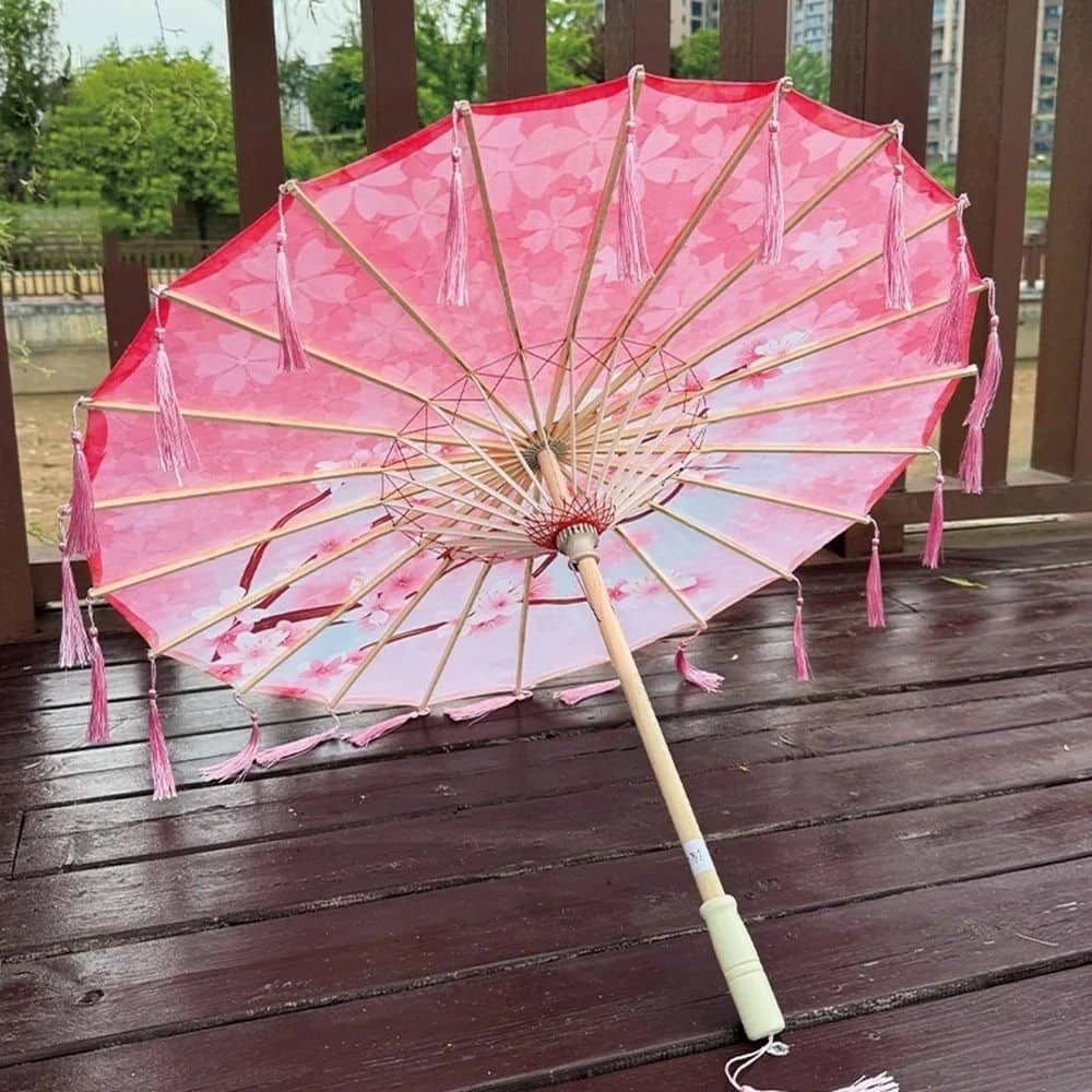 Ombrelle pour femme anti-UV rose avec motifs floraux, décorée de franges, posée sur un sol en bois