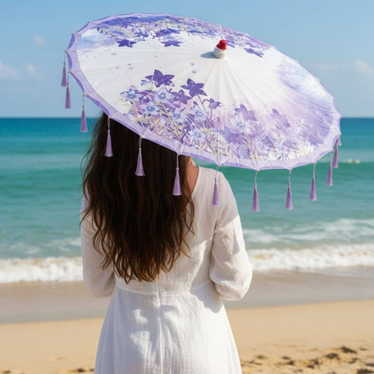 Femme de dos tenant une ombrelle violette ornée de fleurs, sur une plage ensoleillée avec des vagues