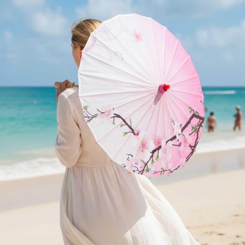 Femme portant une ombrelle rose ornée de fleurs, face à la mer sur une plage ensoleillée