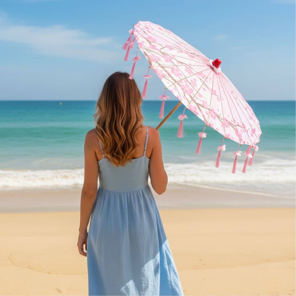 Femme vue de dos sur la plage, tenant une ombrelle rose à motifs floraux sous un ciel bleu