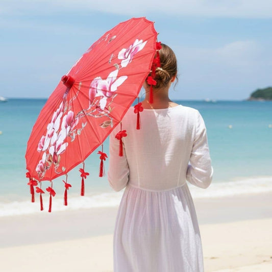Femme en robe blanche tenant une ombrelle rouge ornée de fleurs, face à une plage ensoleillée