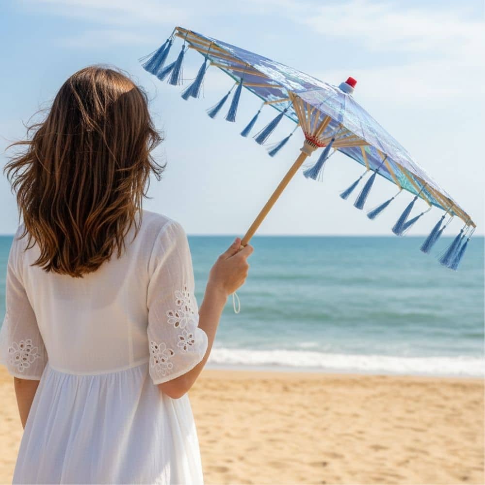 Femme tenant une ombrelle bleue à franges, regardant la mer depuis une plage ensoleillée