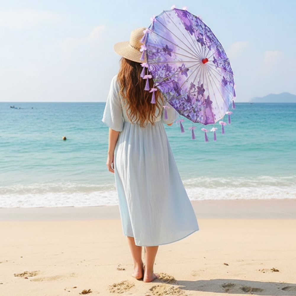 Femme debout sur la plage, portant une robe légère, tenant une ombrelle violette ornée de motifs floraux