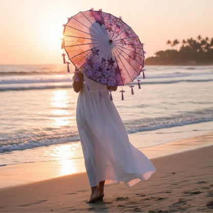 Femme en robe blanche tenant une ombrelle violette ornée de fleurs, marchant sur la plage au coucher du soleil