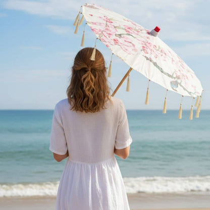 Femme en robe blanche tenant une ombrelle beige et rose, face à la mer sous un ciel bleu
