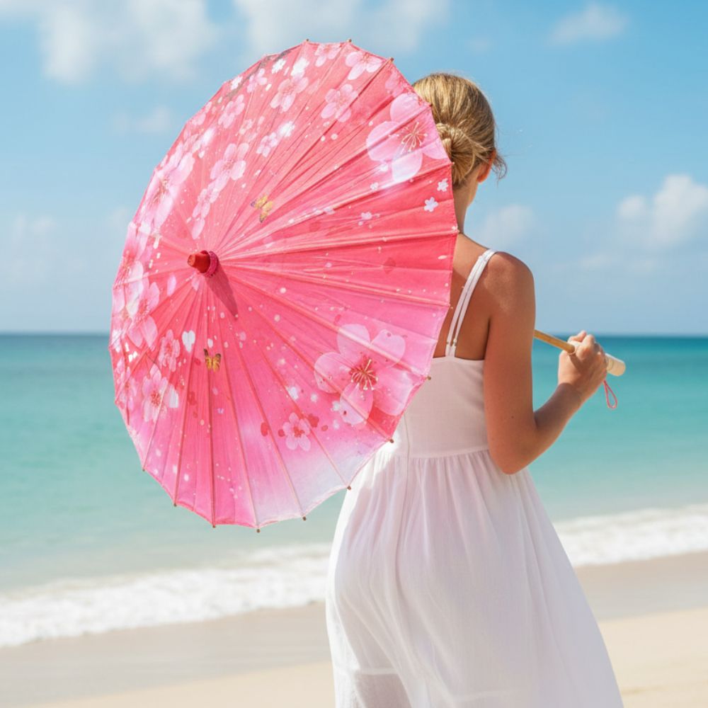 Une ombrelle japonaise en papier rose à motifs floraux, tenue par une femme en robe blanche, sur une plage ensoleillée