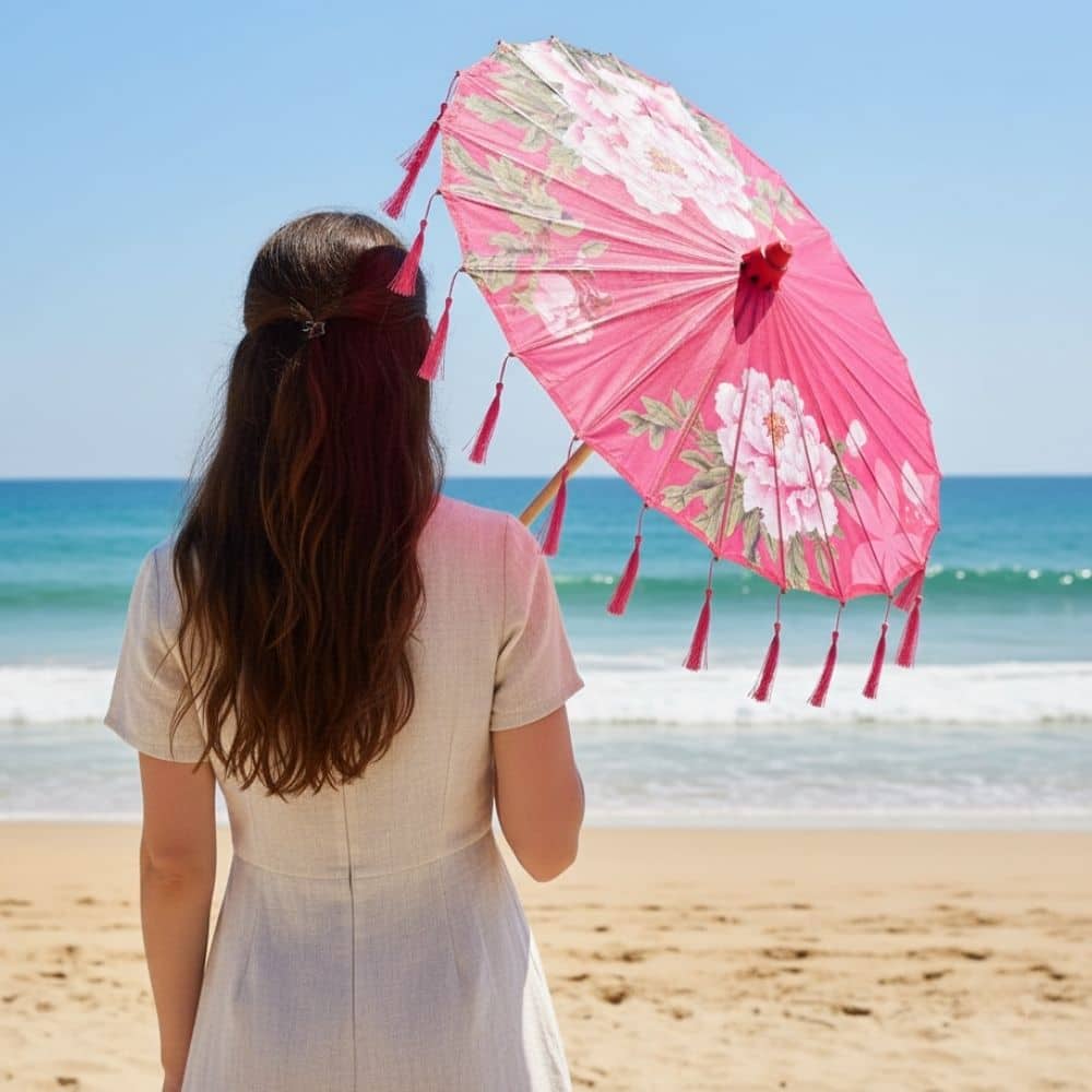 Une femme de dos tient une ombrelle japonaise rose ornée de fleurs, face à la mer et au ciel bleu