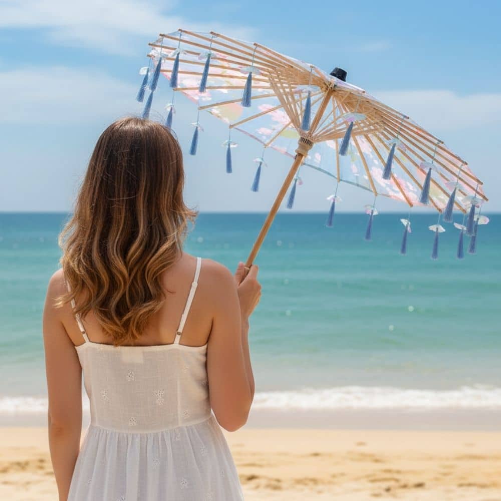 Une femme de dos tient un parapluie japonais bleu décoré de pompons, face à la mer et au ciel clair