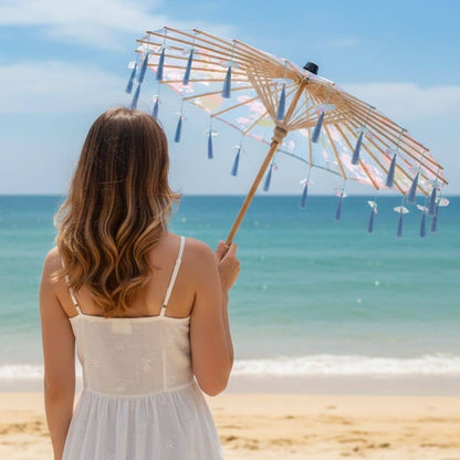 Une femme de dos tient un parapluie japonais bleu décoré de pompons, face à la mer et au ciel clair