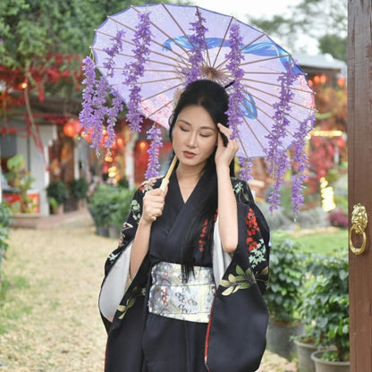 Femme en kimono noir tenant un parapluie violet décoré de fleurs, dans un jardin avec des lanternes