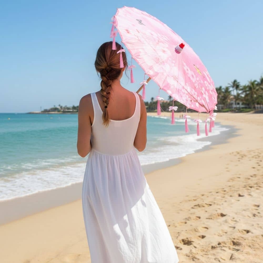 Femme en robe blanche tenant un parapluie japonais rose à pompons sur une plage ensoleillée
