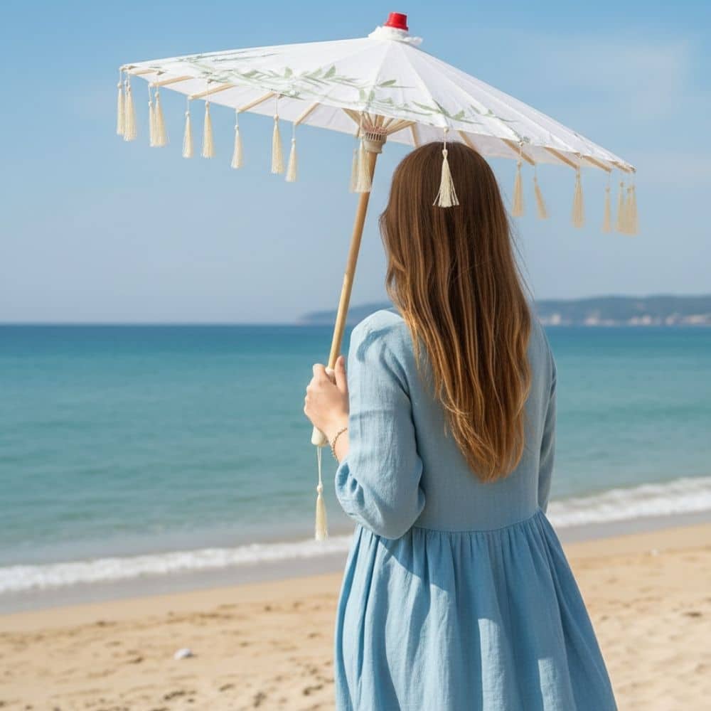 Une femme en robe bleu clair tient un parapluie japonais blanc sur une plage ensoleillée, face à la mer