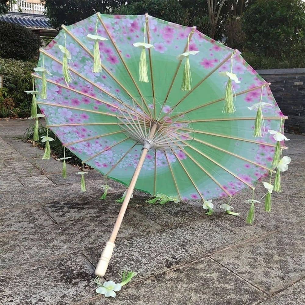 Parapluie ombrelle anti-UV vert décoré de fleurs roses et de franges vertes, posé sur un sol en pierre