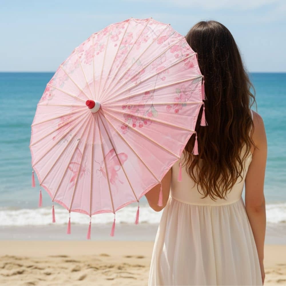 Une femme tient un parapluie rose décoré de motifs floraux, face à la mer et au sable