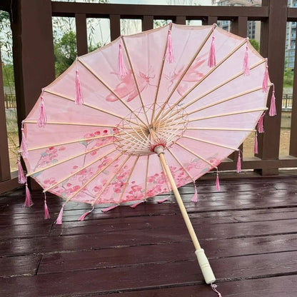 Parapluie UV rose avec motifs floraux, tiges en bois et franges décoratives, posé sur un sol en bois