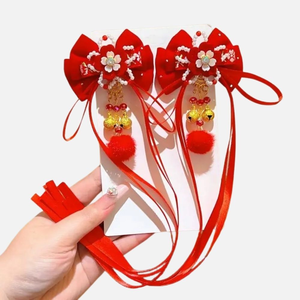Deux accessoires rouges en satin avec des fleurs blanches, pompons et perles dorées, tenus dans une main