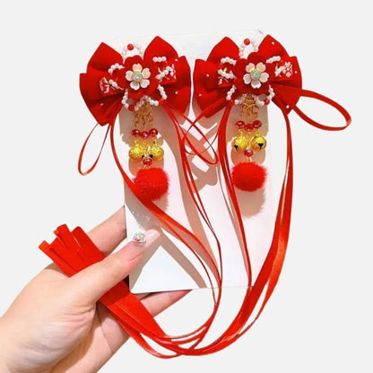 Deux accessoires rouges en satin avec des fleurs blanches, pompons et perles dorées, tenus dans une main