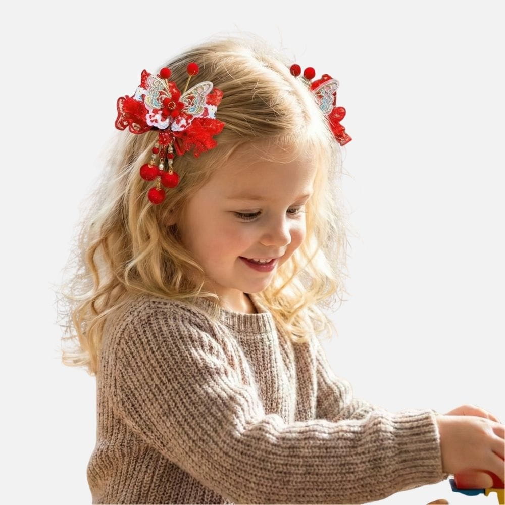 Une petite fille souriante avec des cheveux bouclés porte un pull beige et des accessoires rouges dans les cheveux