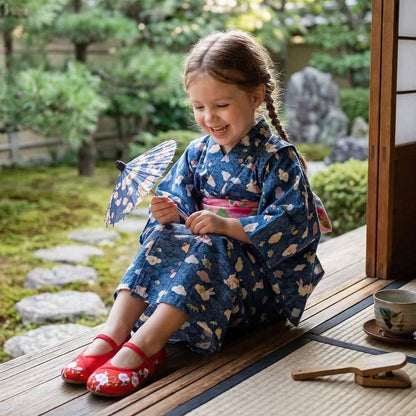 Une petite fille souriante en kimono bleu avec des motifs, portant des chaussures rouges, assise dans un jardin japonais