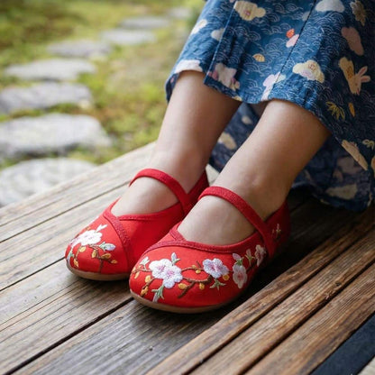 Des ballerines rouges ornées de fleurs brodées, posées sur un sol en bois, avec une jupe à motifs bleus