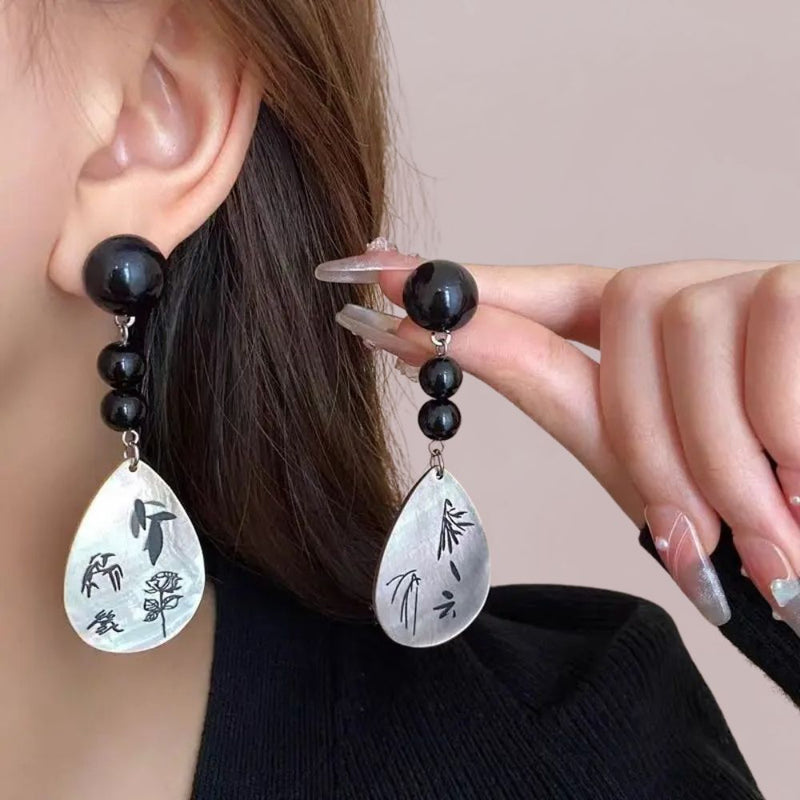 Boucles d'oreilles en nacre avec motifs floraux, perles noires brillantes, portées par une femme en pull noir, style japonais.