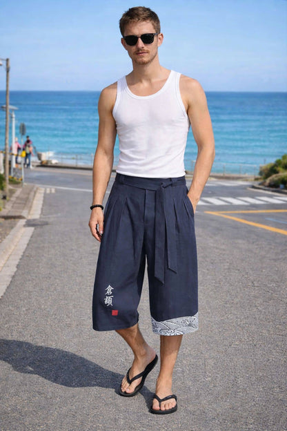 Homme portant un hakama bleu foncé, décoré de motifs blancs, dans une posture décontractée, en arrière plan, une rue et la mer.