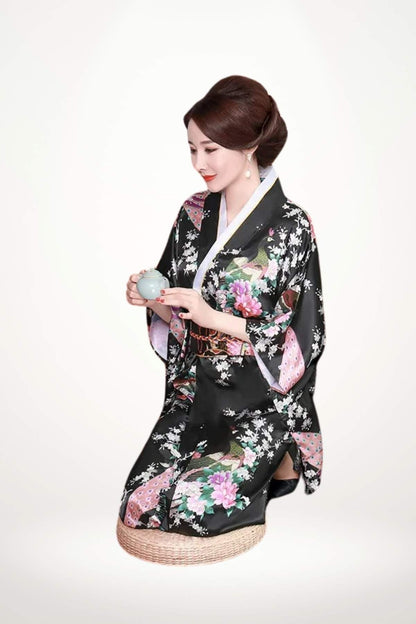 Femme assise en kimono noir à motifs floraux colorés, tenant une tasse, sur un fond neutre