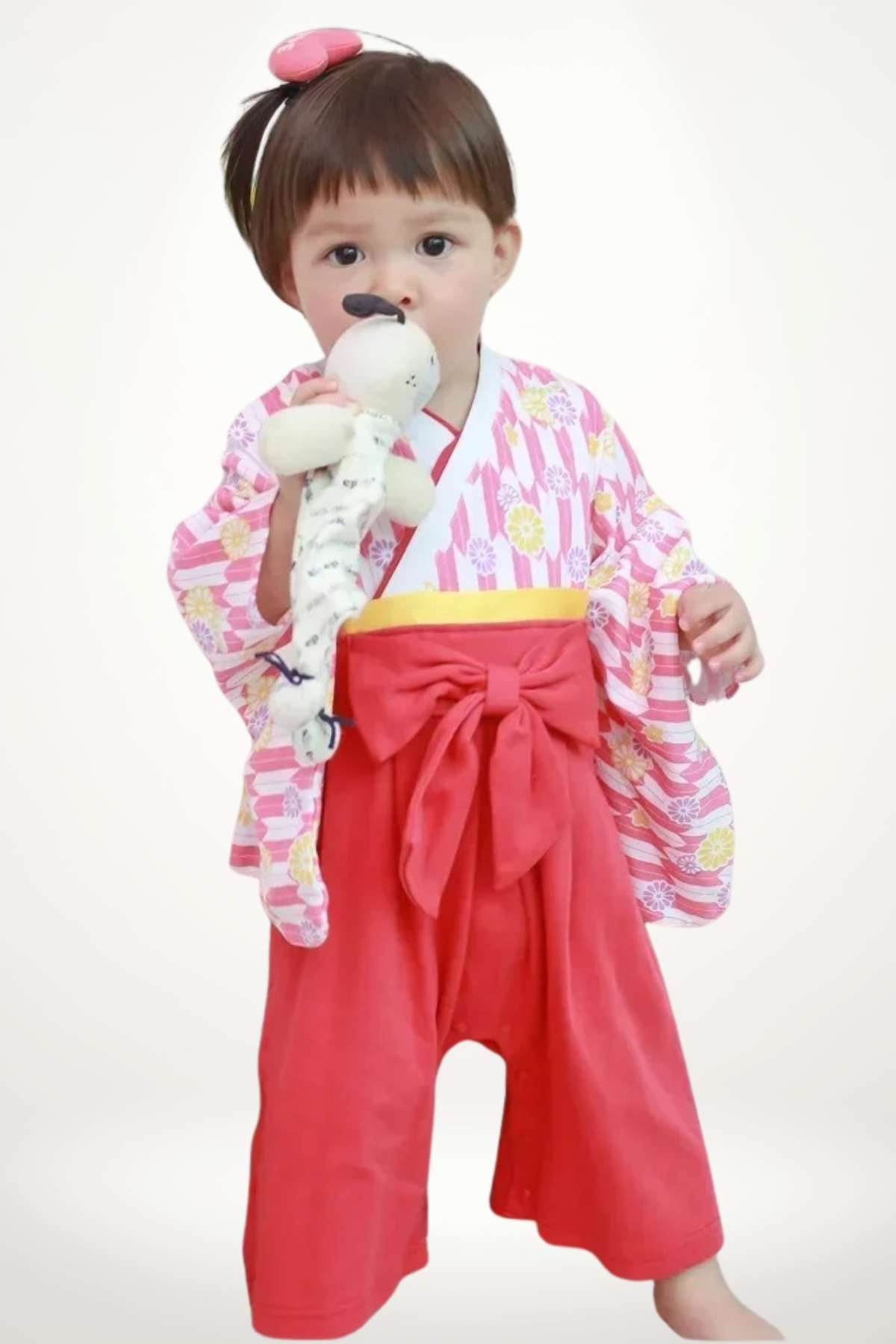 Enfant portant un kimono rose à motifs floraux et un pantalon rouge avec un nœud, tenant une peluche